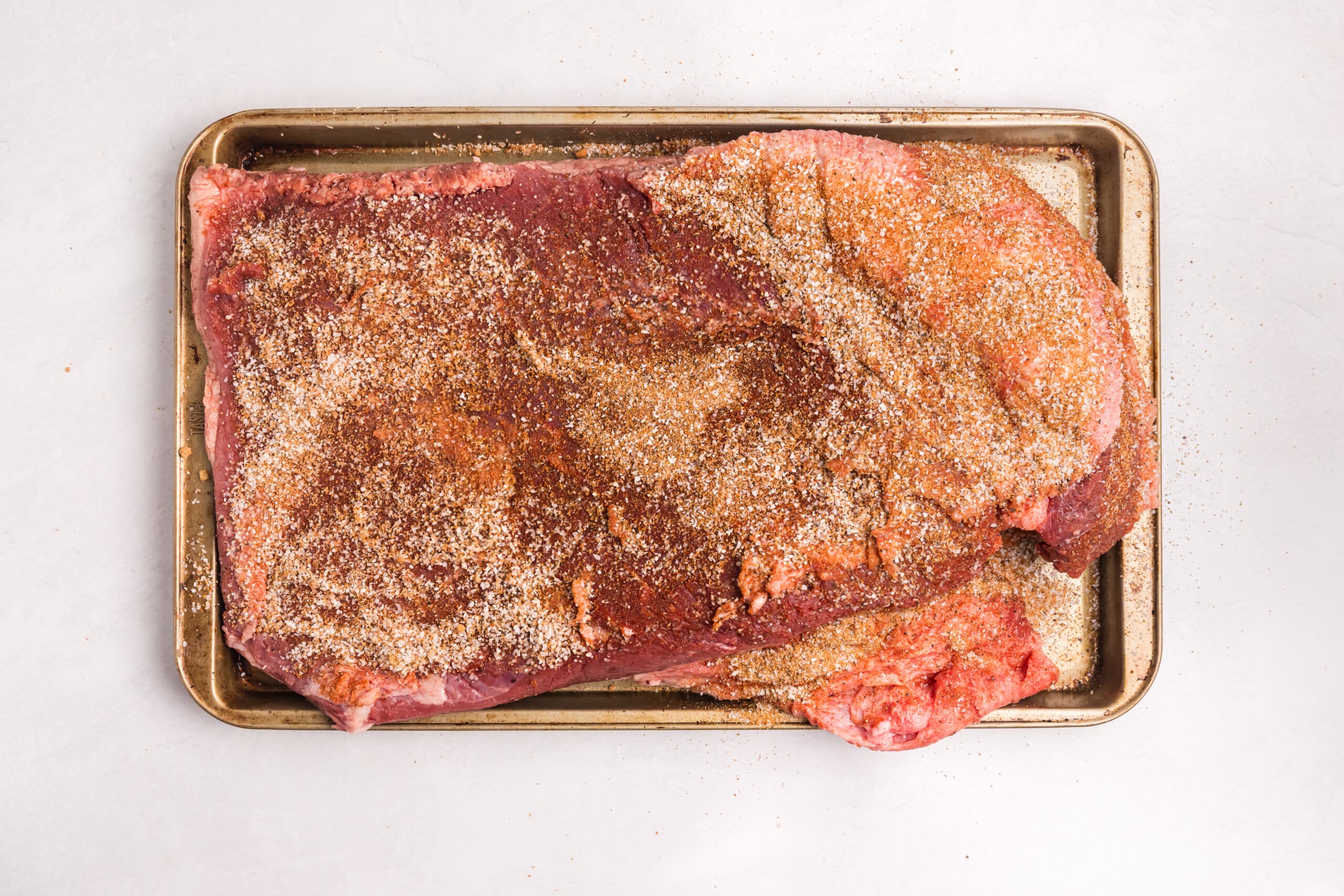 Raw beef brisket generously coated with dry rub seasoning on a baking sheet.
